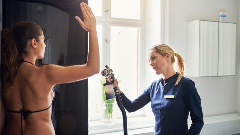 A woman receiving a spray tan in a well-lit room, showcasing a healthy glow and a relaxed atmosphere.